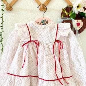 May include: A white, long-sleeved dress with small red polka dots. The dress features red ribbon accents, including bows, and a ruffled collar and sleeves. The dress is hanging on a pink hanger with a cat illustration.
