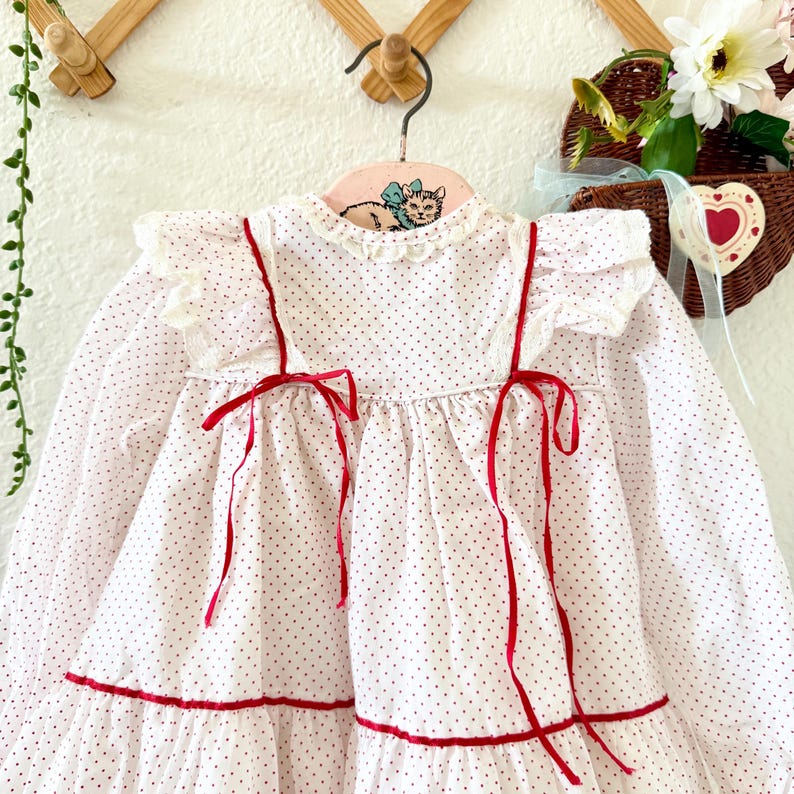 May include: A white, long-sleeved dress with small red polka dots. The dress features red ribbon accents, including bows, and a ruffled collar and sleeves. The dress is hanging on a pink hanger with a cat illustration.