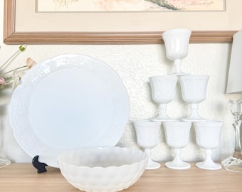 May include: A set of white vintage tableware, featuring a large plate with a grape pattern, a textured bowl, and a stack of stemmed goblets. The items are displayed on a wooden surface, with a painting in the background.