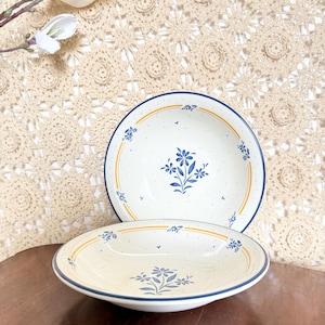 May include: Two off-white ceramic bowls with blue floral designs. The bowls have a speckled texture, blue rims, and yellow accent lines. The bowls are on a wooden surface with a crocheted background.