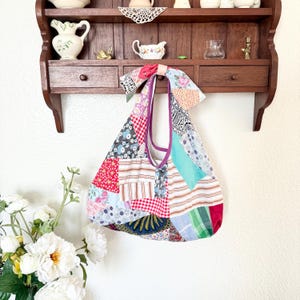 May include: A hobo-style shoulder bag constructed from a patchwork of various fabric patterns, including red gingham, floral prints, stripes, and solid teal. The bag features a knotted top handle and a purple interior lining visible at the opening.