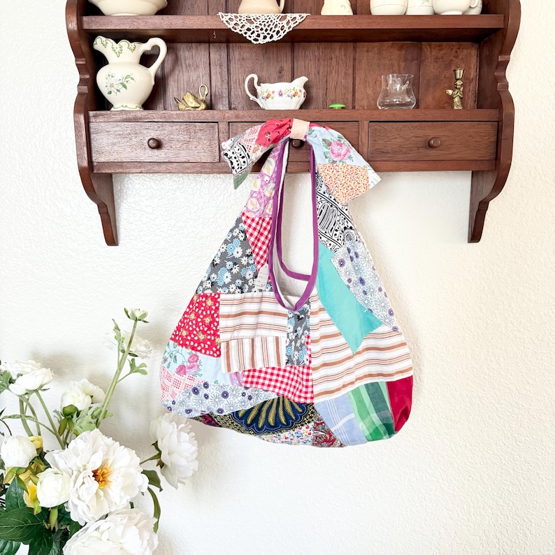 May include: A hobo-style shoulder bag constructed from a patchwork of various fabric patterns, including red gingham, floral prints, stripes, and solid teal. The bag features a knotted top handle and a purple interior lining visible at the opening.