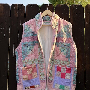 May include: A sleeveless quilted vest featuring a patchwork design of various floral and solid fabric squares in shades of pink, blue, lavender, and green. The garment has a stand-up collar, two front patch pockets, and a solid white interior lining.
