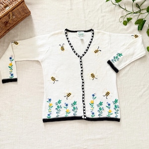 May include: A white long-sleeve knit cardigan featuring embroidered yellow and black bees, blue and yellow flowers along the hem and sleeves, and a black and white checkered trim along the V-neckline and button-down front closure.