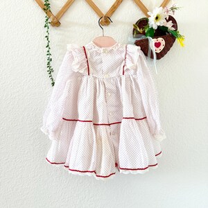 May include: A white and red polka dot dress with long sleeves and a ruffled collar. The dress has red trim and buttons down the front. The dress is hanging on a wooden hanger against a white wall.