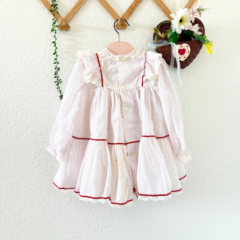 May include: A white and red polka dot dress with long sleeves and a ruffled collar. The dress has red trim and buttons down the front. The dress is hanging on a wooden hanger against a white wall.