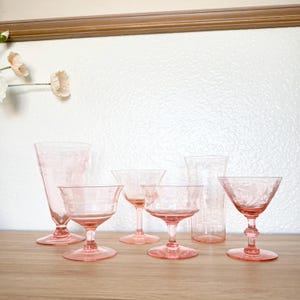 Könnte beinhalten: Eine Sammlung rosa Glaswaren, darunter Sektflöten, Coupés und eine hohe Vase, auf einer Holzoberfläche angeordnet. Die Gläser haben geätzte Blumenmuster und sind in verschiedenen Formen und Größen erhältlich, ideal für eine Bar im Vintage-Stil.