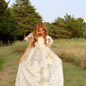 May include: A floor-length cream-colored dress constructed from various panels of lace and embroidered fabric. The garment features short ruffled sleeves, a square neckline, and a patchwork design incorporating floral motifs in yellow and red.
