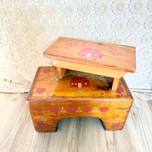 以下が含まれることがあります： 2つの木製ステップスツール。上段のスツールにはピンク色の「WELCOME」の文字と赤い家のグラフィックが描かれています。下段のスツールには赤いハートと家のグラフィックがあります。どちらも木材に着色されています。