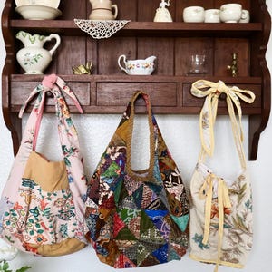 May include: Three fabric bags hanging from a wooden shelf. The left bag is pink with floral patterns and a knotted strap. The center bag features a multi-colored patchwork design with various geometric prints. The right bag is cream with floral motifs and yellow ribbon ties.