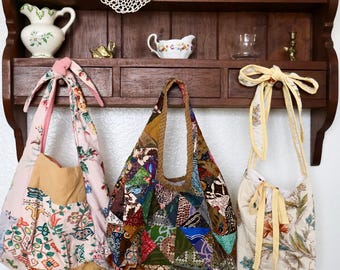 May include: Three fabric bags hanging from a wooden shelf. The left bag is pink with floral patterns and a knotted strap. The centre bag features a multi-coloured patchwork design with various geometric prints. The right bag is cream with floral motifs and yellow ribbon ties.