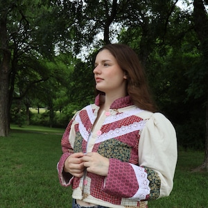 May include: A cropped quilted jacket featuring a patchwork design of red, green, and cream floral and plaid fabrics. The jacket has a high collar, puff sleeves, white eyelet lace trim along the chest, and three pink buttons down the front.