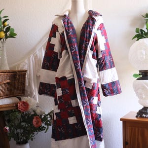 May include: A long, open-front coat constructed from quilted fabric featuring a patchwork pattern of solid white squares alternating with dark blue and red floral print squares. The garment has a wide collar and long sleeves.