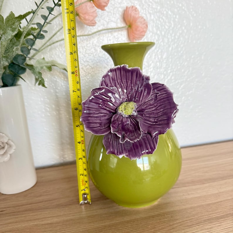 May include: A green ceramic vase with a large, purple flower accent. The vase is approximately 8 inches tall. The flower has a yellow center. The vase is on a wooden surface.