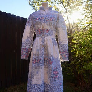 May include: A long-sleeved, button-front coat constructed from quilted fabric featuring a patchwork design. The pattern consists of white, light blue, and pink floral prints combined with geometric star motifs throughout the garment.