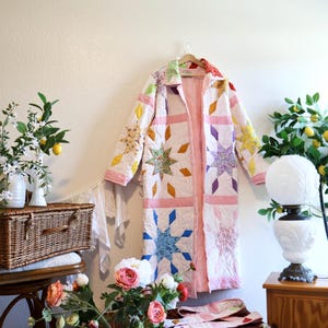 May include: A long, quilted coat featuring a patchwork design of multi-colored star patterns on a light pink background. The garment has a collared neckline, long sleeves, and a button-down front, constructed from various floral and solid fabric pieces.
