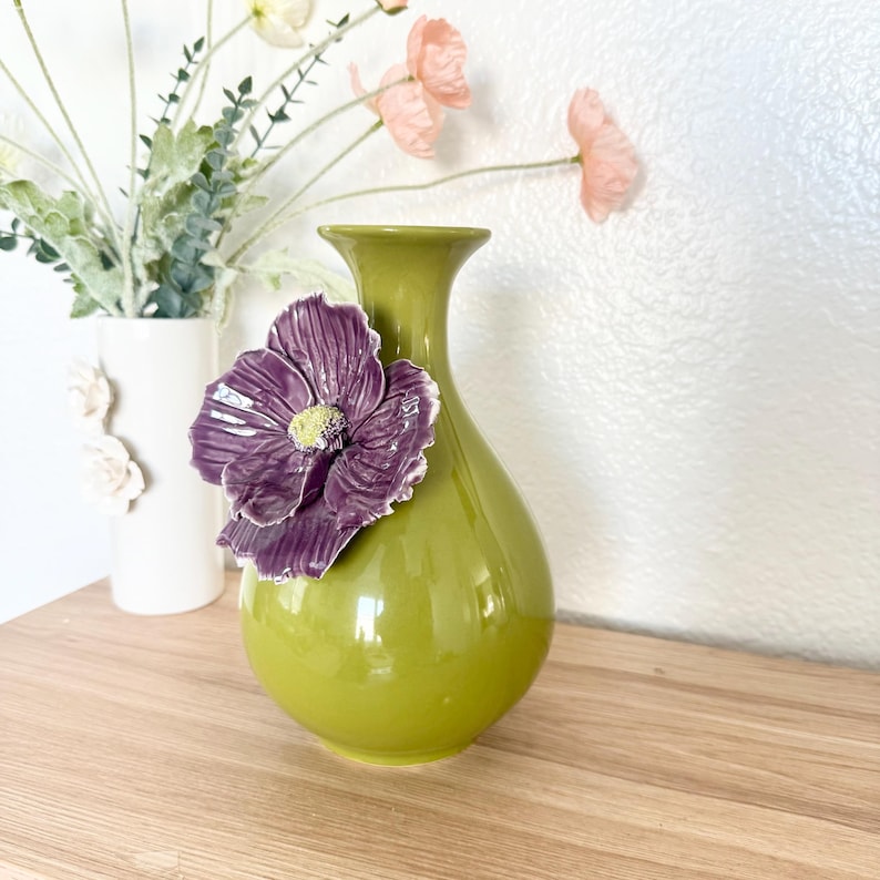 May include: A green ceramic vase with a large, purple flower accent. The flower has a yellow center and is attached to the front of the vase. A white vase with floral accents and faux flowers are in the background.