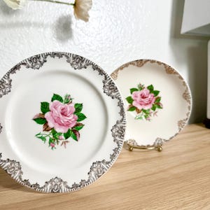 Pode incluir: Dois pratos decorativos vintage. Um é branco com uma rosa rosa e folhas verdes, e uma borda preta e dourada ornamentada. O outro é branco sujo com uma rosa rosa e uma borda dourada.