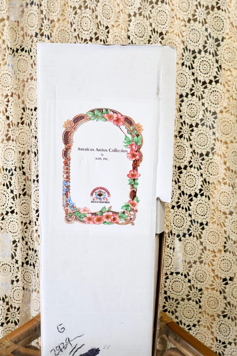 May include: A tall white rectangular cardboard box featuring a decorative label with a floral border. The label text reads American Artists Collection by Kais, Inc. and World Gallery Dolls & Collectibles. The letter G and number 2929 are written in black ink.