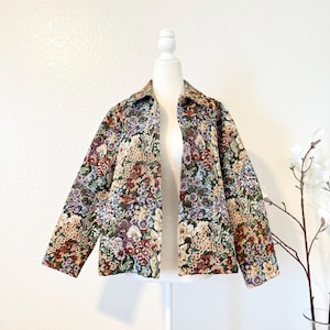May include: A floral tapestry jacket with a collared neckline and long sleeves. The jacket features a colorful pattern of flowers in shades of purple, brown, green, and cream. It is displayed on a white mannequin.