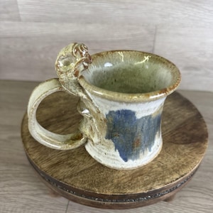 May include: Handmade ceramic mug with a unique sculpted handle. The mug features a cream-colored base with blue and brown accents. The rim and handle have a textured, organic design. The mug is displayed on a small, round wooden stand.