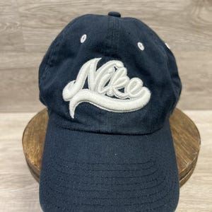 May include: Navy blue baseball cap with a curved brim. The cap features the white Nike logo embroidered on the front. The hat is made of a durable fabric and has a classic, adjustable design.
