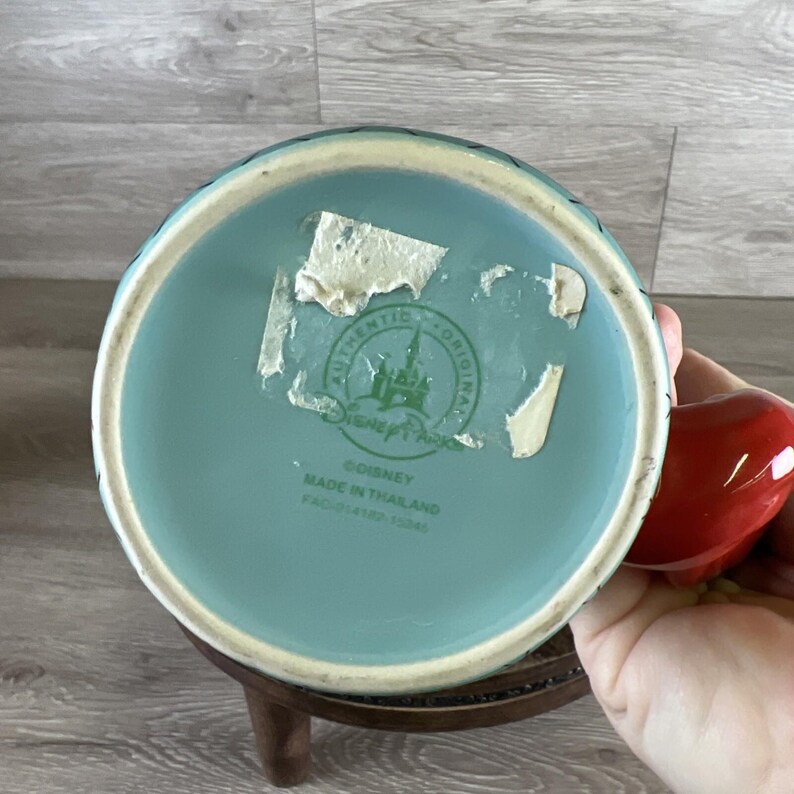 May include: A light blue ceramic mug with a red apple-shaped handle. The bottom of the mug has a light blue glaze and a partially visible sticker that reads "Disney Park Authentic Original". The mug is made in Thailand.