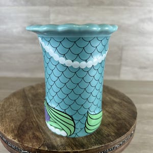 May include: A ceramic vase with a mermaid scale design. The vase is light blue with black scale outlines, a white pearl necklace design, and green and purple mermaid tail details. The top edge has a scalloped design.