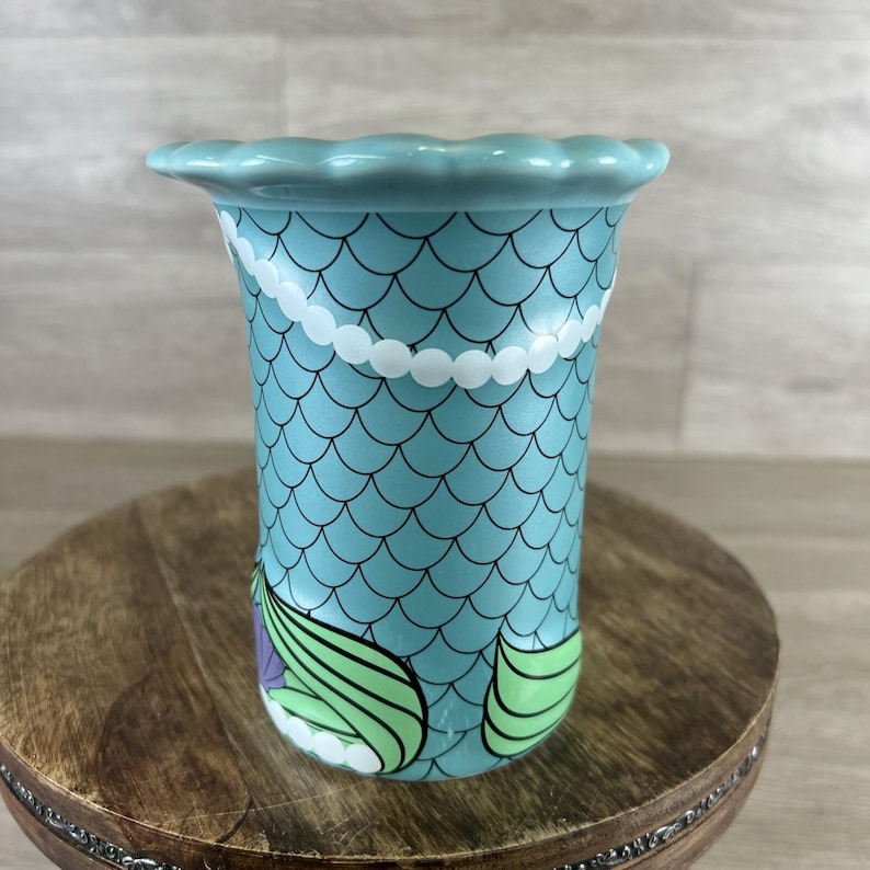 May include: A ceramic vase with a mermaid scale design. The vase is light blue with black scale outlines, a white pearl necklace design, and green and purple mermaid tail details. The top edge has a scalloped design.
