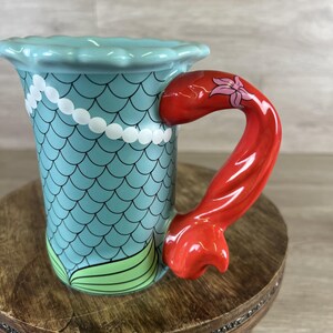May include: A ceramic mug with a mermaid theme. The mug features a light blue body with a scale pattern and a red tail-shaped handle. A white pearl necklace design adorns the mug, and green fins are at the base. The handle has a pink flower design.
