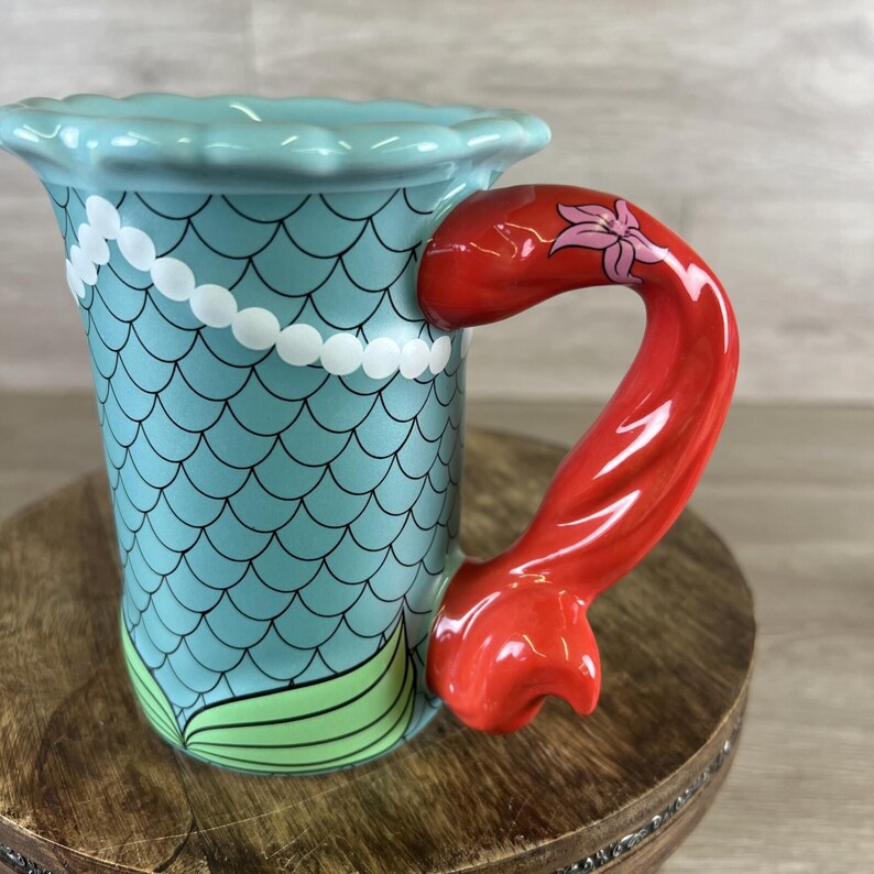May include: A ceramic mug with a mermaid theme. The mug features a light blue body with a scale pattern and a red tail-shaped handle. A white pearl necklace design adorns the mug, and green fins are at the base. The handle has a pink flower design.
