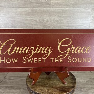 May include: A rectangular, burgundy-colored wooden sign with the words "Amazing Grace" and "How Sweet the Sound" in gold script. The sign is displayed on a wooden stand, suitable for home decor.