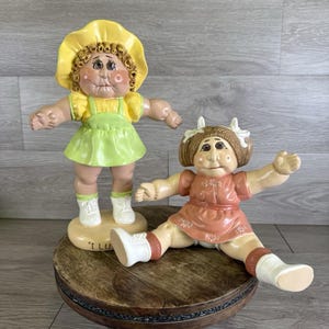 May include: Two vintage Cabbage Patch Kids dolls. One doll is standing and wearing a yellow hat, green dress, and white shoes. The other doll is sitting and wearing a peach dress and white socks. Both dolls have brown hair and rosy cheeks.