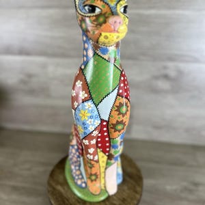 May include: A colorful, patchwork-style cat figurine. The cat is painted with various patterns and colors, including blue, green, red, and orange. It is sitting on a small, round wooden stand. The cat has a pink nose and detailed facial features.