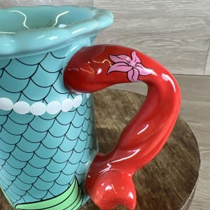 May include: A ceramic mug with a mermaid theme. The mug features a turquoise body with a scale pattern and a red handle shaped like a mermaid tail. A pink flower decorates the handle. The mug has a white beaded trim.