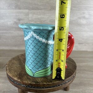May include: A ceramic mermaid-themed mug with a blue scale pattern, white pearl accents, and a red handle. The mug is approximately 5 inches tall, measured with a yellow tape measure. The mug sits on a small wooden stand.