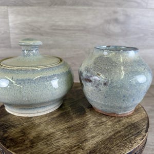 May include: Two ceramic pieces: a lidded jar and a small vase. The jar is round with a lid and a small knob. The vase is a rounded shape with a wide opening. Both pieces have a speckled, blue-green glaze and sit on a wooden surface.