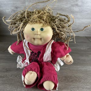 May include: A vintage doll with a round face, green eyes, and light brown yarn hair. The doll wears a pink shirt with a graphic, pink pants, and a white dotted bib. The doll's hands and feet are made of a soft, beige material.
