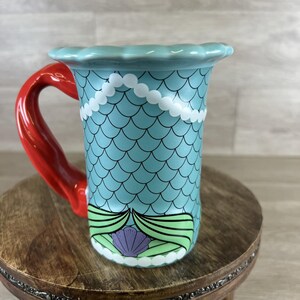 May include: A ceramic mug with a mermaid theme. The mug features a light blue body with a scale pattern, a red handle, and a scalloped rim. White pearl-like accents and a seashell design complete the look.