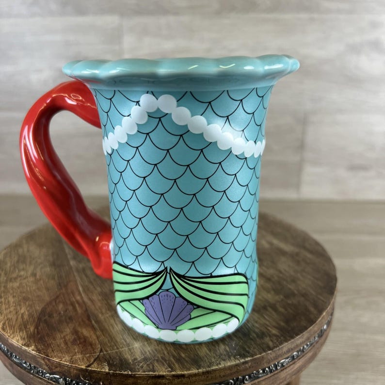 May include: A ceramic mug with a mermaid theme. The mug features a light blue body with a scale pattern, a red handle, and a scalloped rim. White pearl-like accents and a seashell design complete the look.