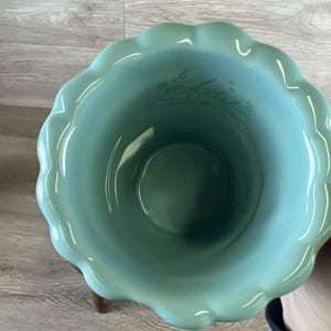 May include: A teal ceramic bowl with a scalloped rim. The interior features the name "Ariel" in a cursive font. The bowl is set on a wooden stand, showcasing a decorative and functional design.