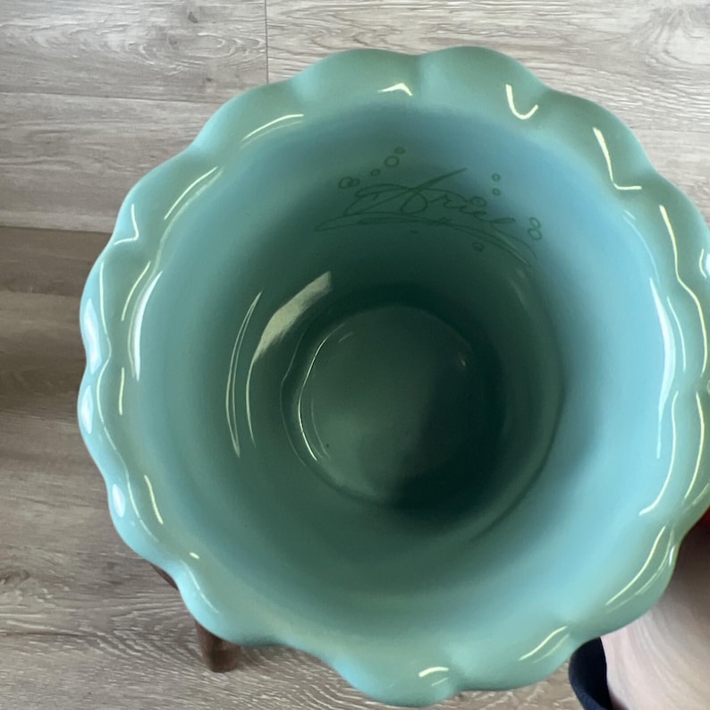 May include: A teal ceramic bowl with a scalloped rim. The interior features the name "Ariel" in a cursive font. The bowl is set on a wooden stand, showcasing a decorative and functional design.