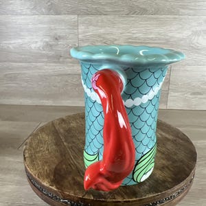 May include: A ceramic mermaid-themed pitcher with a turquoise body featuring a scale pattern. The handle is a vibrant red mermaid tail. The top rim is scalloped and light blue. The pitcher is decorated with white and green accents.