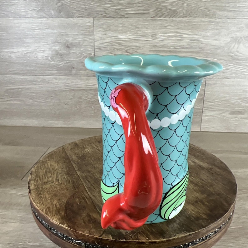 May include: A ceramic mermaid-themed pitcher with a turquoise body featuring a scale pattern. The handle is a vibrant red mermaid tail. The top rim is scalloped and light blue. The pitcher is decorated with white and green accents.