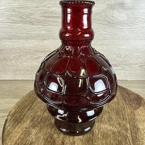 May include: A ruby red glass vase with a textured, geometric pattern. The vase has a bulbous body and a cylindrical neck with a decorative rim. The vase is displayed on a wooden surface.