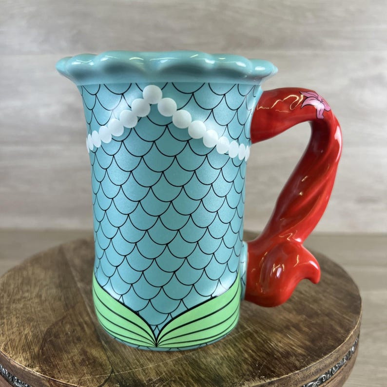 May include: A ceramic mermaid-themed mug. The mug features a light blue body with a scale pattern and a white pearl necklace design. The handle is a vibrant red mermaid tail. The base has green seaweed accents.