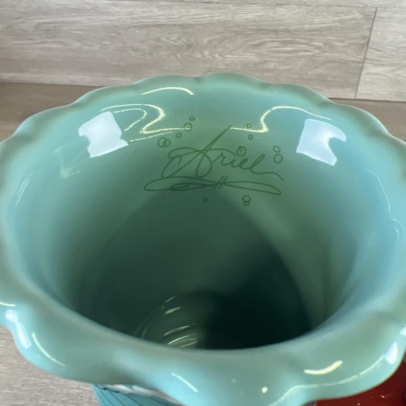 May include: A teal ceramic vase with a scalloped rim. The word "Ariel" is written in a cursive font on the inside of the vase. The vase has a wide opening and a rounded body.