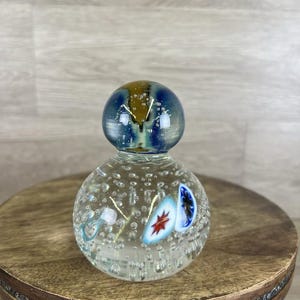May include: A clear glass decorative bottle with a round body and a spherical stopper. The bottle is filled with tiny bubbles and has colorful, abstract designs. The stopper is blue and gold. The bottle sits on a small, round wooden table.