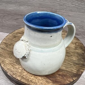 May include: A ceramic mug with a rounded body and a wide handle. The exterior is off-white with a speckled finish, while the interior and rim are a deep blue. A textured, white, shell-shaped decorative element is attached to the side of the mug.