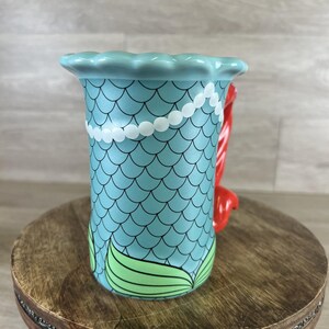 May include: A ceramic mug with a mermaid theme. The mug features a light blue scale pattern, a white pearl necklace design, and a red handle. Green mermaid tail fins are at the base of the mug.